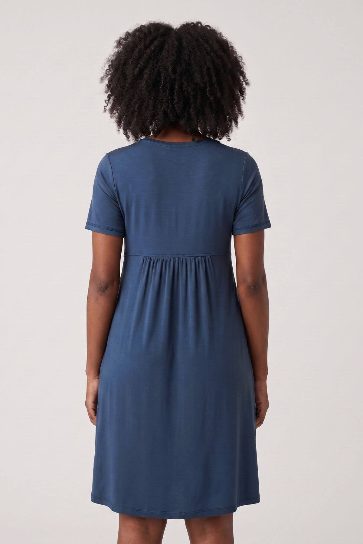 A woman viewed from the back wearing a short-sleeve blue dress with a gathered waist seam, set against a plain light gray studio background.