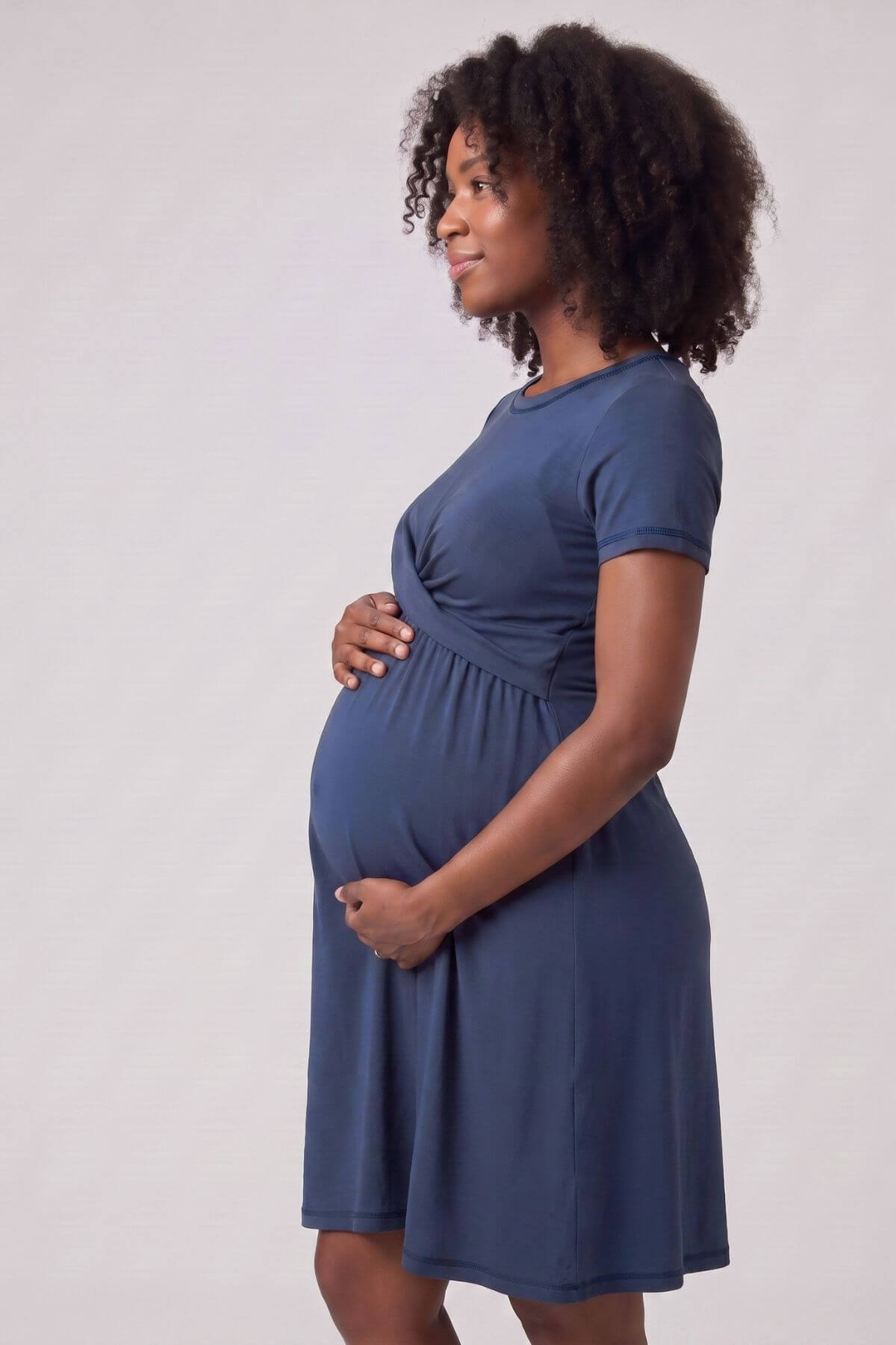 A pregnant woman with curly black hair is shown in profile wearing a short-sleeve, knee-length blue maternity dress against a plain light gray studio background.