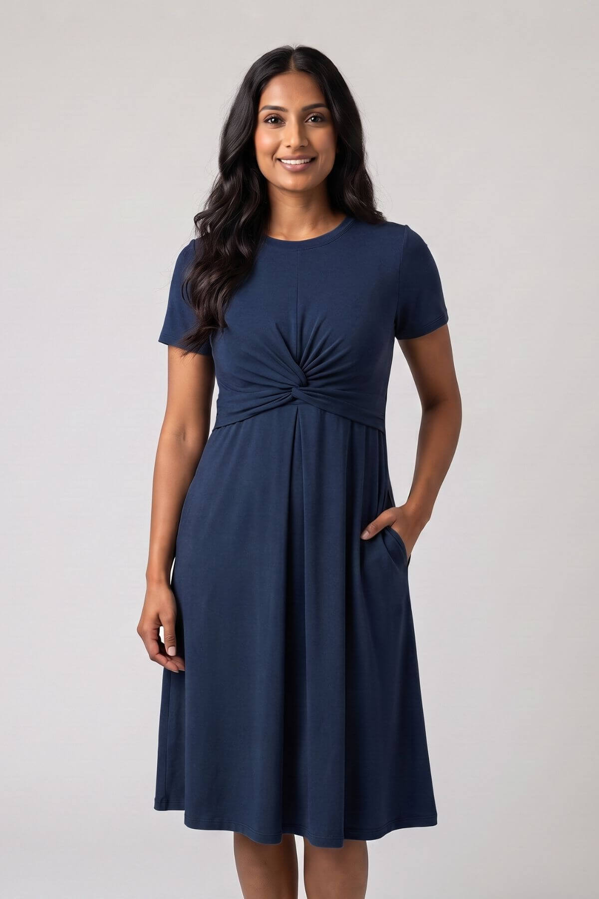A woman modeling a navy blue short-sleeve A-line dress with a gathered twist detail at the waist and side pockets, set against a plain light gray studio background.