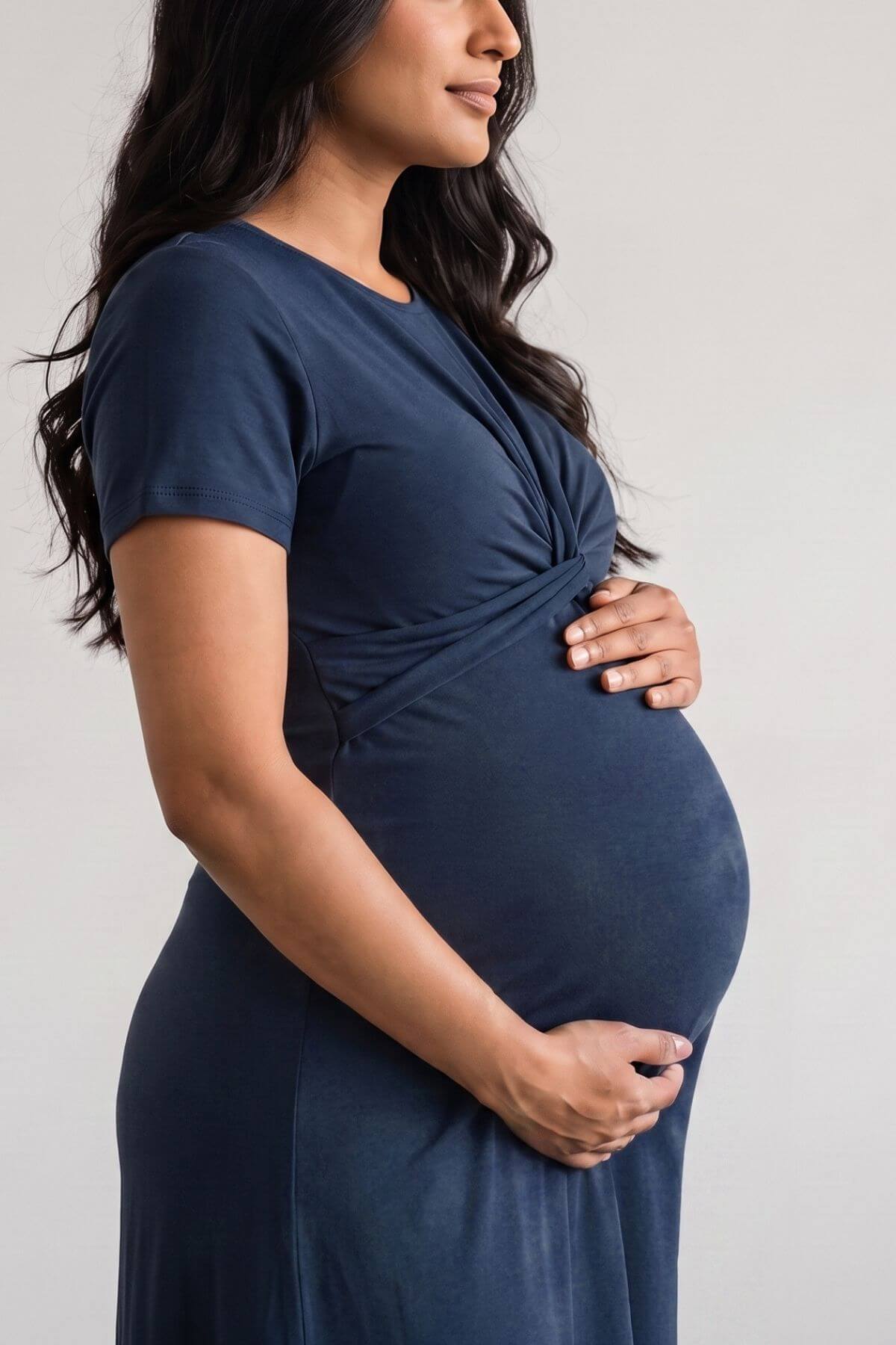 A pregnant woman wearing a navy blue short-sleeve maternity dress with a twisted front detail, shown from the side against a light gray background.