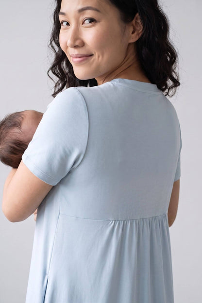 A woman wearing a light blue short-sleeve maternity dress, holding a baby against her chest, shown from the back against a light gray background.