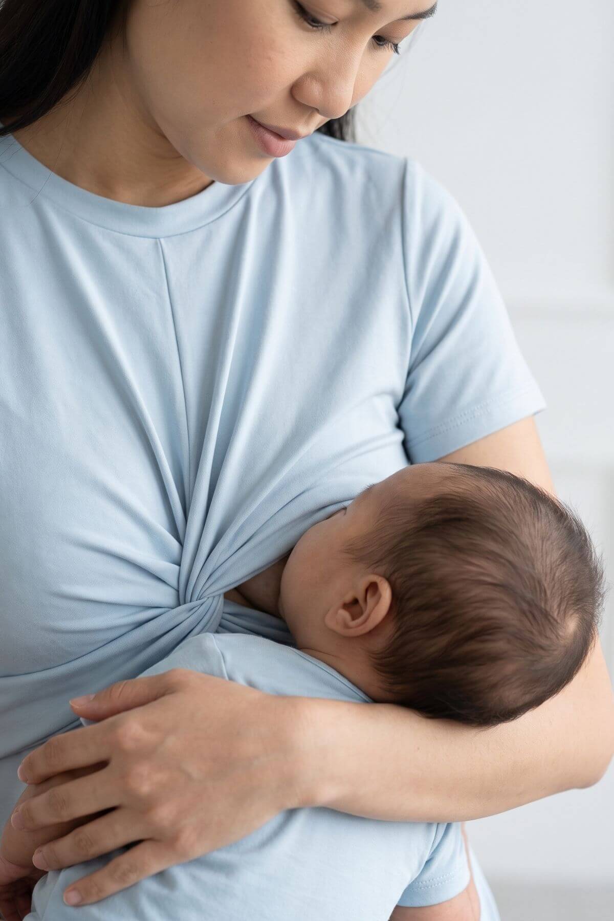 A close-up lifestyle shot of a woman breastfeeding a baby, wearing a light blue t-shirt with the fabric twisted to create a pouch.