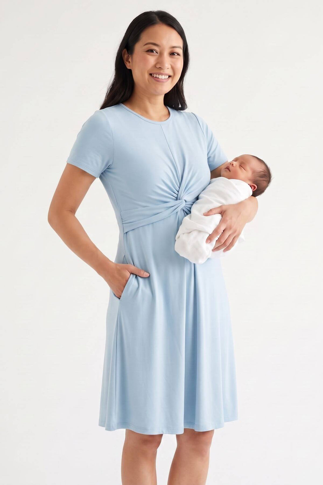 A woman wearing a light blue short-sleeve maternity dress with a twisted waist and side pockets, holding a sleeping newborn baby wrapped in a white swaddle, against a plain white studio background.