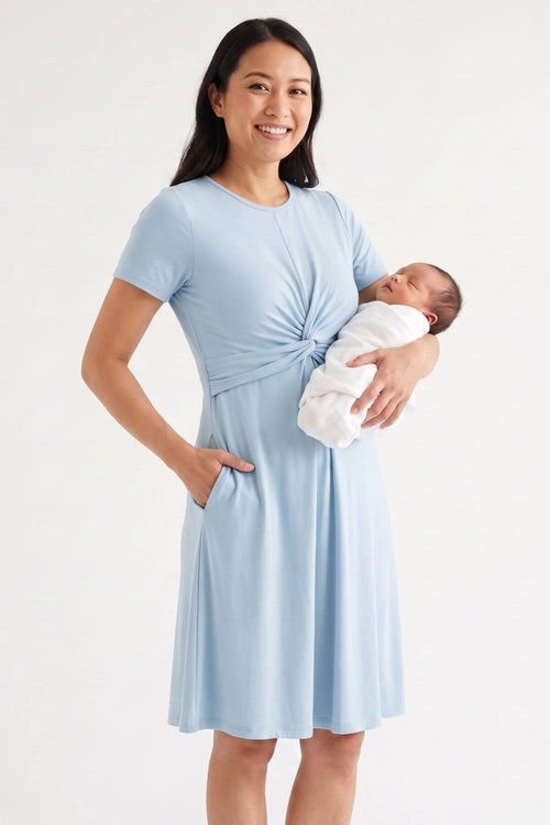 A woman wearing a light blue short-sleeve maternity dress with a twisted waist and side pockets, holding a sleeping newborn baby wrapped in a white swaddle, against a plain white studio background.#color_ether