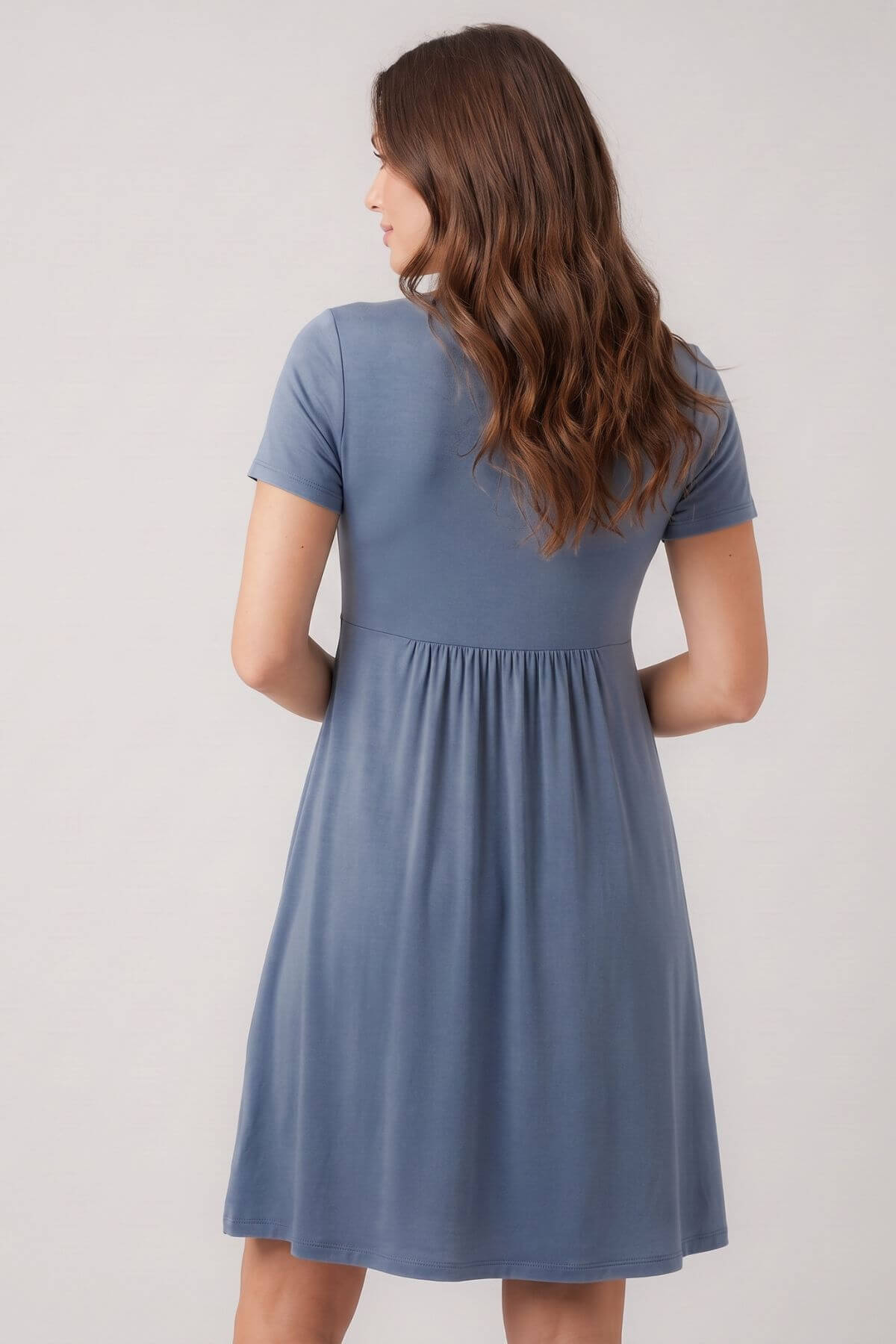 A woman viewed from the back wearing a short-sleeve blue dress with a gathered waist, against a plain light gray studio background.