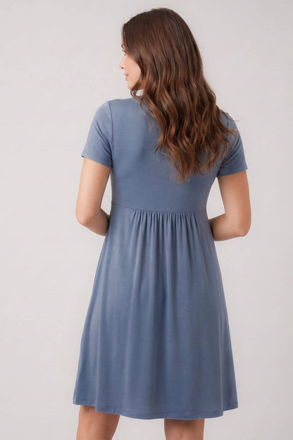 A woman viewed from the back wearing a short-sleeve blue dress with a gathered waist, against a plain light gray studio background.