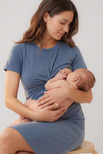 A woman wearing a dusty blue maternity nursing top and matching skirt holds and breastfeeds a baby, seated on a light wood stool against a plain light gray background.