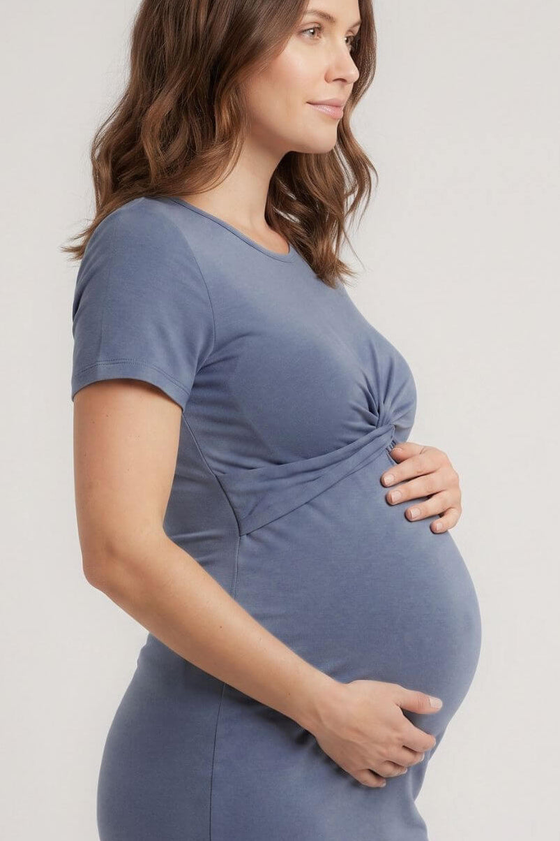 A pregnant woman wearing a slate blue maternity t-shirt dress, standing in profile against a light gray studio background.