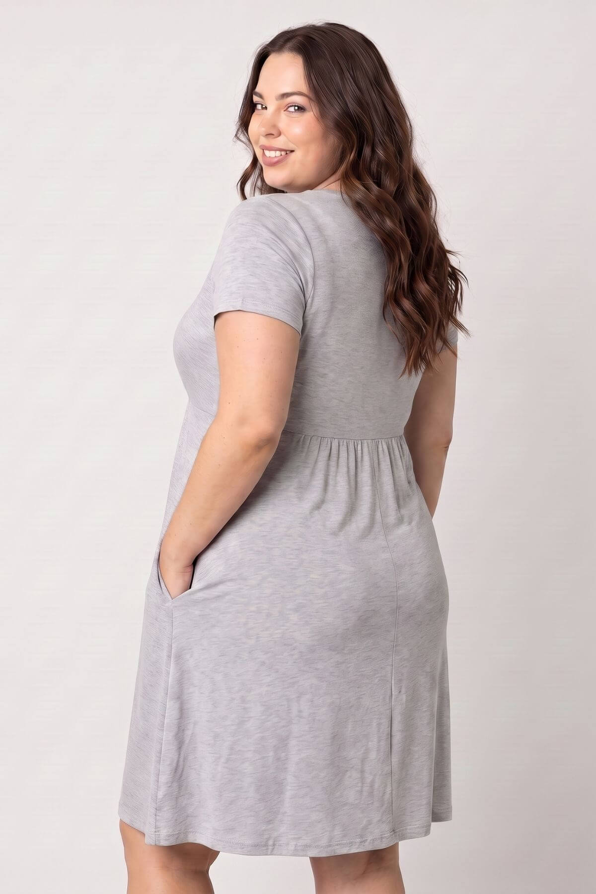 A woman modeling a light gray short-sleeve shift dress with side pockets, shown from a three-quarter back angle against a plain light gray studio background.
