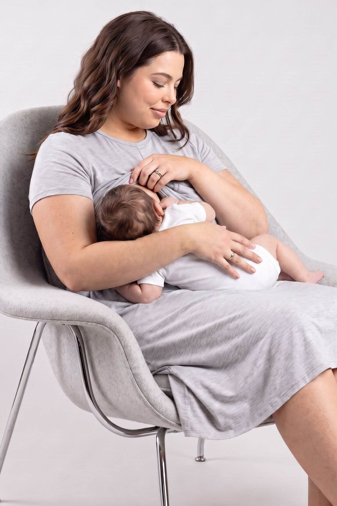 A woman in a light gray t-shirt and matching dress sits in a modern gray chair, breastfeeding a baby dressed in white. Shot in a studio setting with a light gray background.