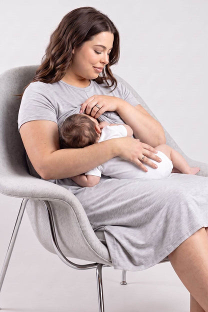 A woman in a light gray t-shirt and matching dress sits in a modern gray chair, breastfeeding a baby dressed in white. Shot in a studio setting with a light gray background.