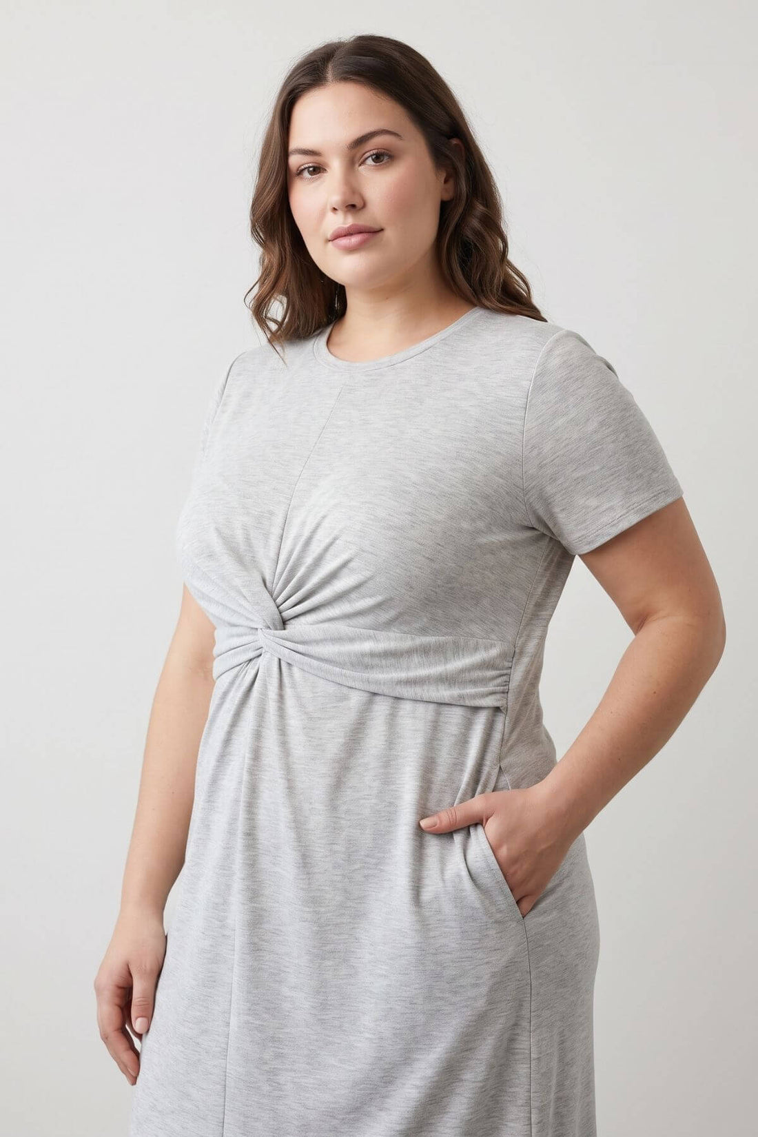 A woman wearing a light gray heather short-sleeve dress with a twisted knot detail at the waist and side pockets, photographed against a plain light gray studio background.