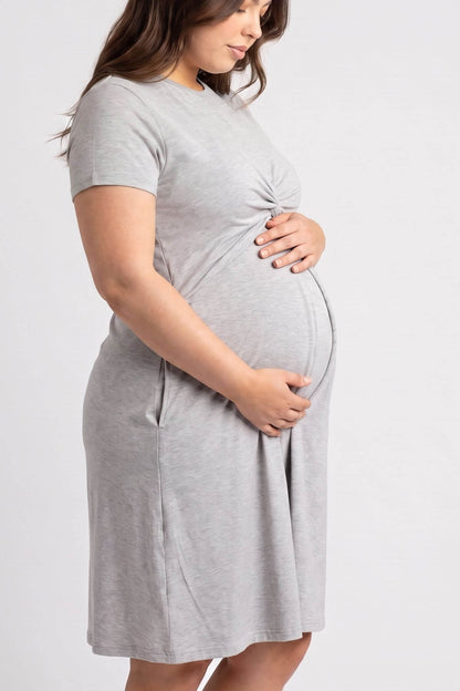 A pregnant woman wearing a light gray short-sleeve maternity dress with side pockets, standing against a plain light gray studio background.