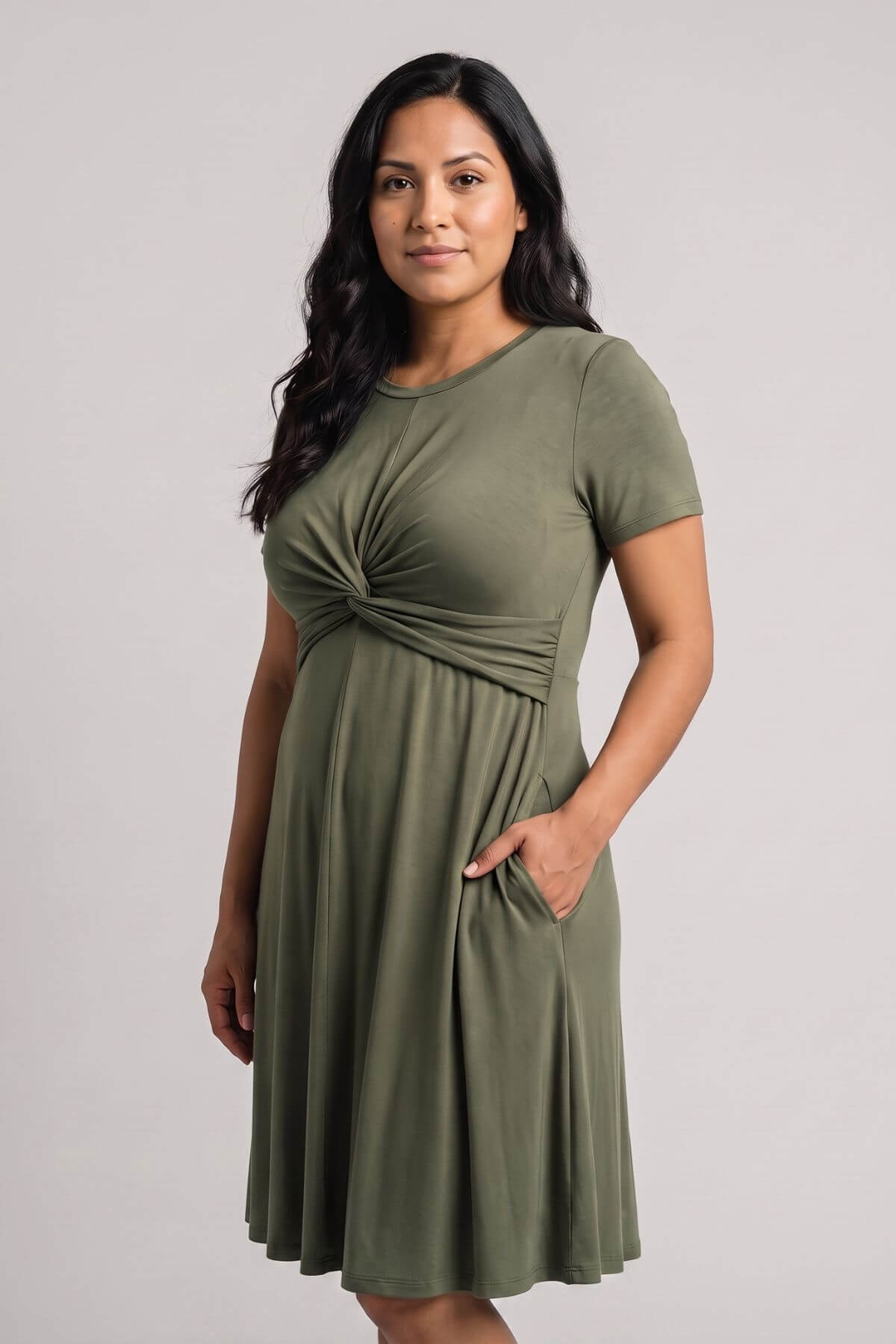A woman modeling a short-sleeve olive green maternity dress with a gathered twist detail at the waist and side pockets, set against a plain light gray studio background.