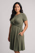 A woman modeling a short-sleeve olive green maternity dress with a gathered twist detail at the waist and side pockets, set against a plain light gray studio background.