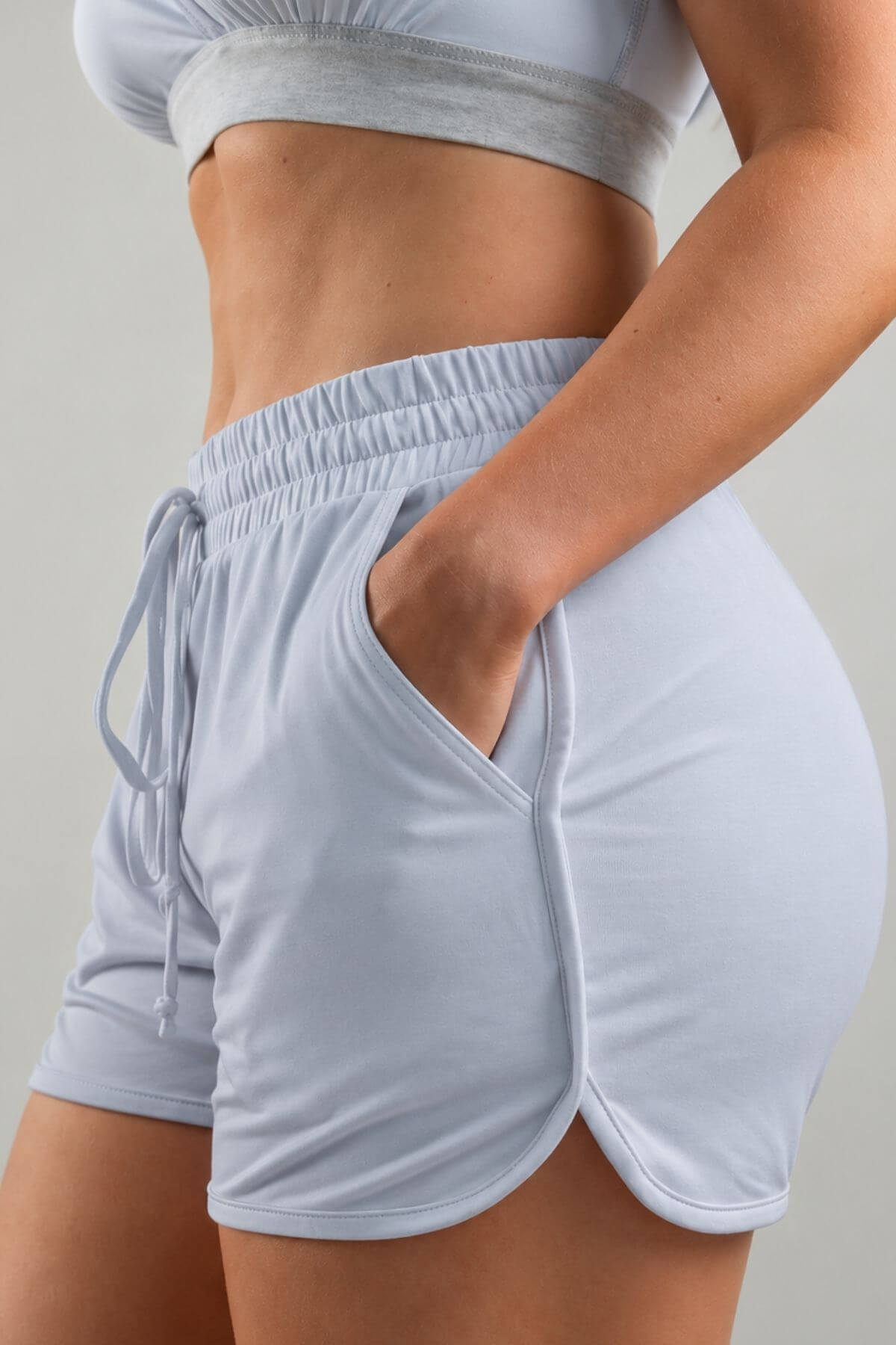 A close-up of a model wearing light blue-gray athletic shorts with side pockets and a drawstring waistband, paired with a matching sports bra against a neutral studio background.