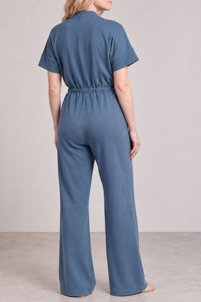 A woman viewed from the back wearing a slate blue short-sleeve jumpsuit with a high waist and wide legs, standing against a neutral studio backdrop..