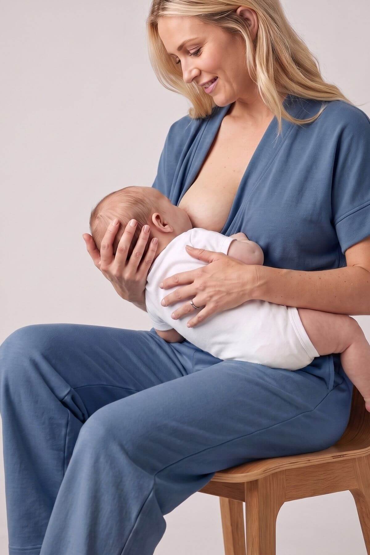 A woman wearing a dusty blue maternity nursing set sits on a wooden stool, holding a baby who is wearing a white onesie. The set consists of a short-sleeve open-front top and matching wide-leg pants, photographed against a neutral light gray background..