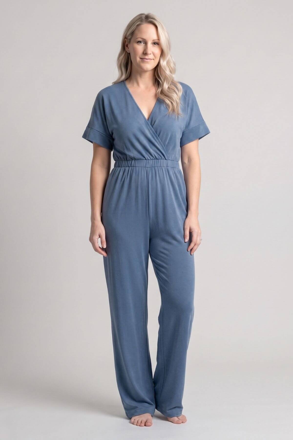 A woman modeling a slate blue short-sleeve wrap jumpsuit against a neutral light gray studio background.