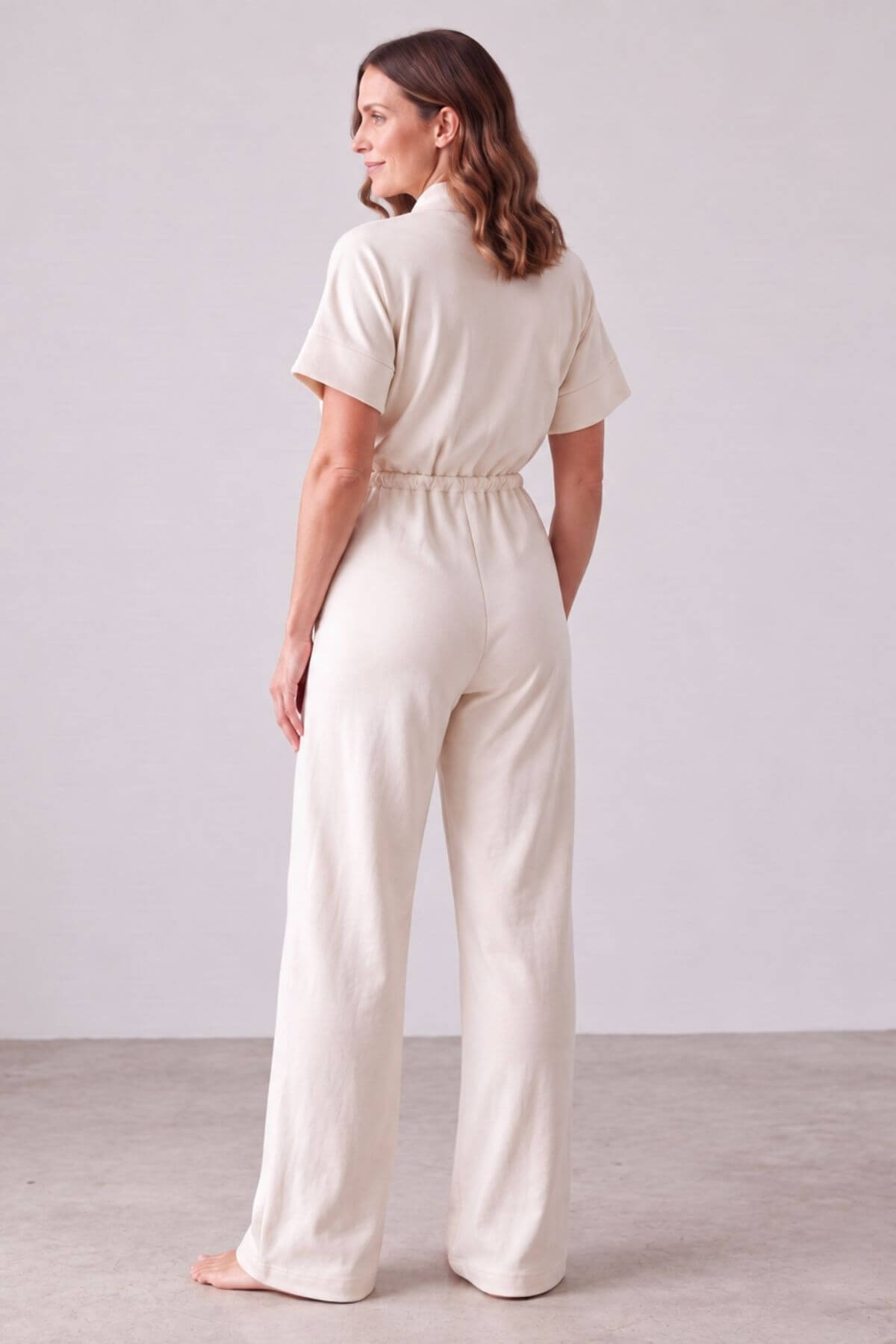 A woman stands barefoot in a studio wearing a cream-colored short-sleeve jumpsuit with a gathered waist and wide legs, shown from the back..