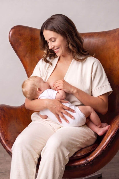 A woman in a cream-colored nursing top and matching pants sits in a brown leather armchair, breastfeeding a baby dressed in a white onesie..