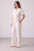 A woman modeling a short-sleeve cream-colored jumpsuit with a wrap-style V-neck and wide-leg pants, standing barefoot against a neutral studio backdrop.