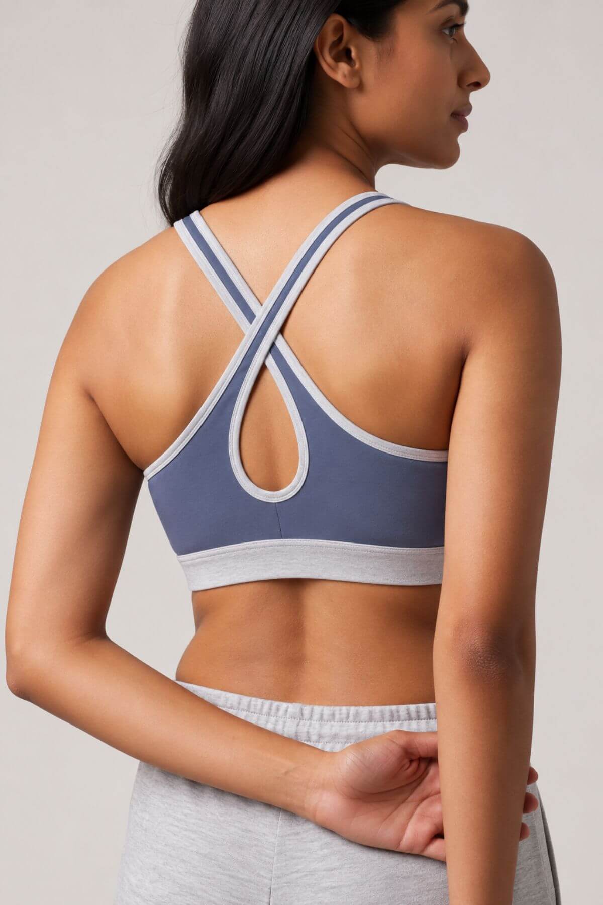 A woman shown from the back wearing a blue-gray sports bra with white trim and a crossover crisscross back design, paired with light gray athletic bottoms, against a plain light gray studio background.