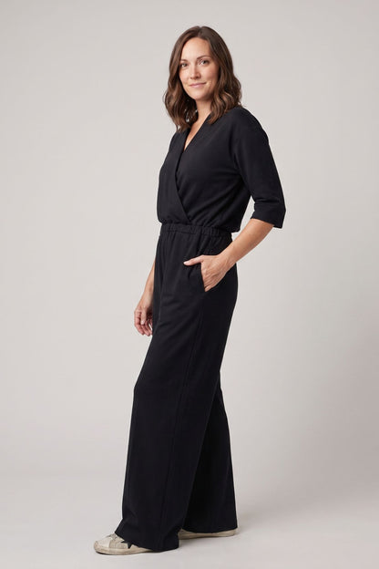 A woman modeling a black wrap-style jumpsuit with three-quarter sleeves and wide-leg pants, standing against a plain light gray studio background.