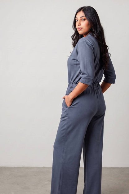 A woman modeling a slate blue jumpsuit, shown from a three-quarters back angle against a neutral studio background.