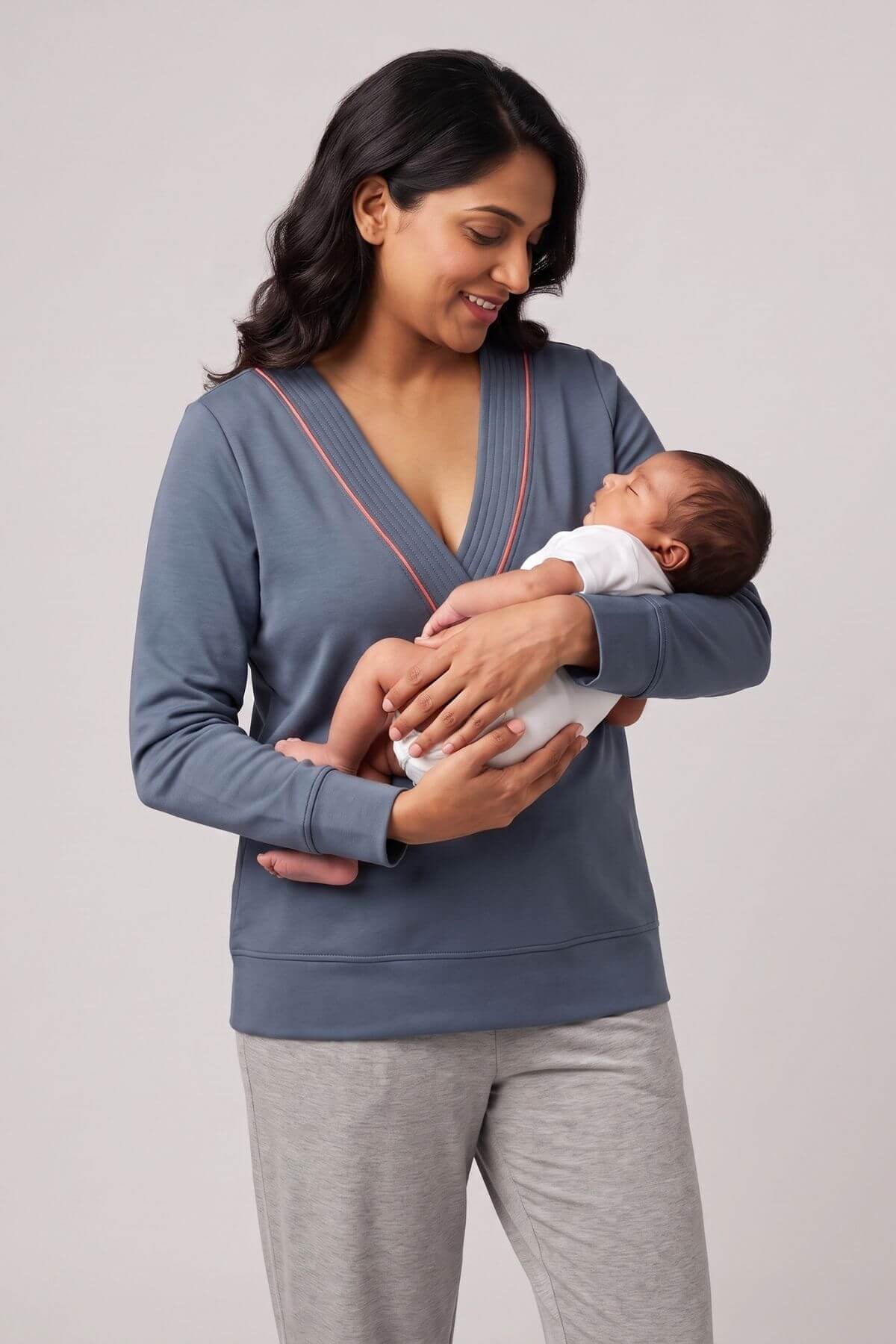 A woman wearing a slate blue long-sleeve maternity nursing top and light gray sweatpants, holding a baby dressed in a white onesie against a plain light gray studio background.