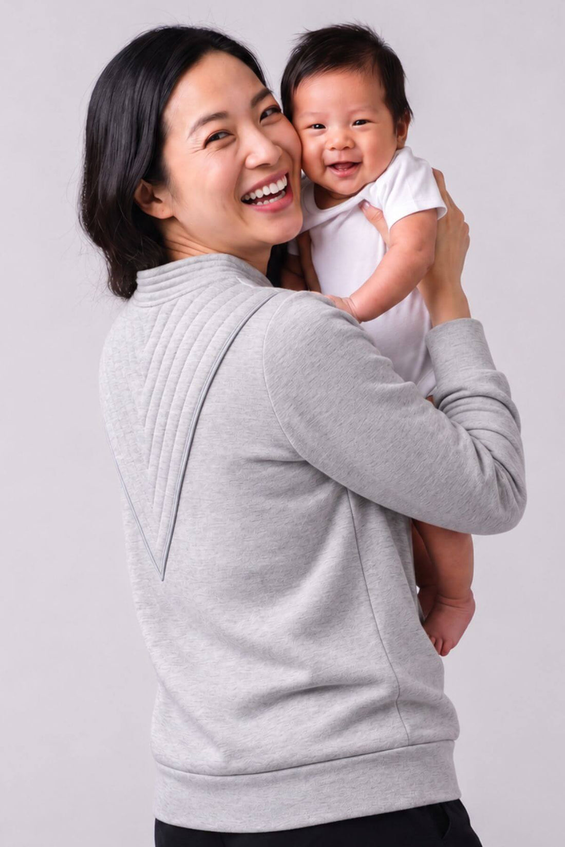 A woman holding a baby, both wearing light gray and white tops respectively, photographed against a plain light gray studio background.