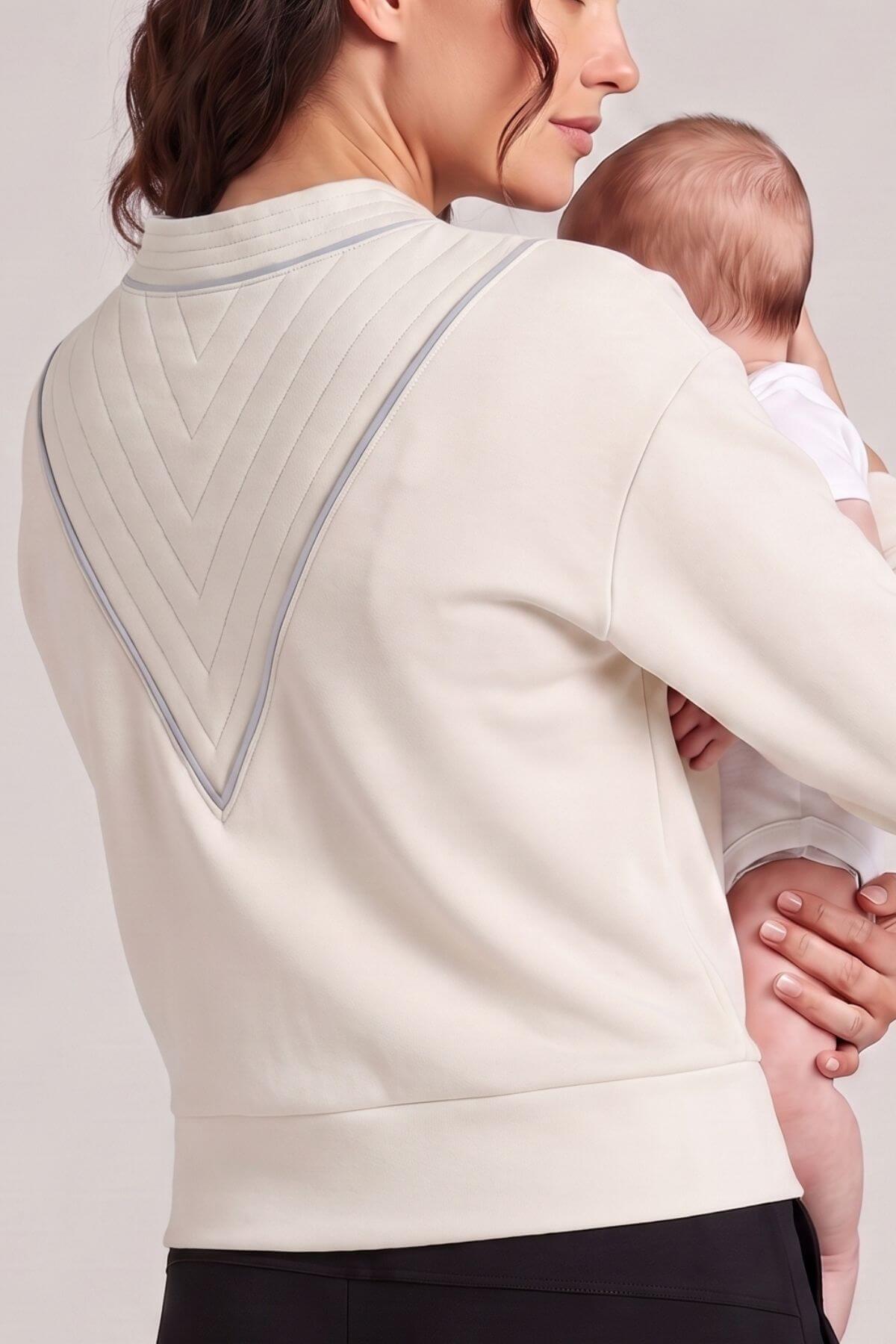 A woman wearing a cream-colored long-sleeve top with a textured chevron pattern on the back, holding a baby against her chest. The photo is cropped from the back of the woman&