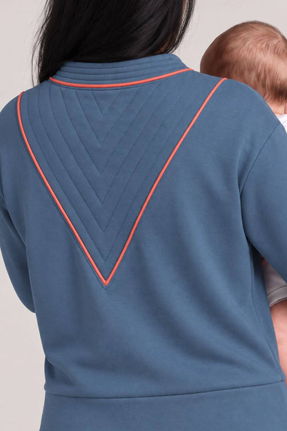 A close-up back view of a person wearing a slate blue sweatshirt with a contrasting orange-red stitched chevron pattern on the back, holding a baby.