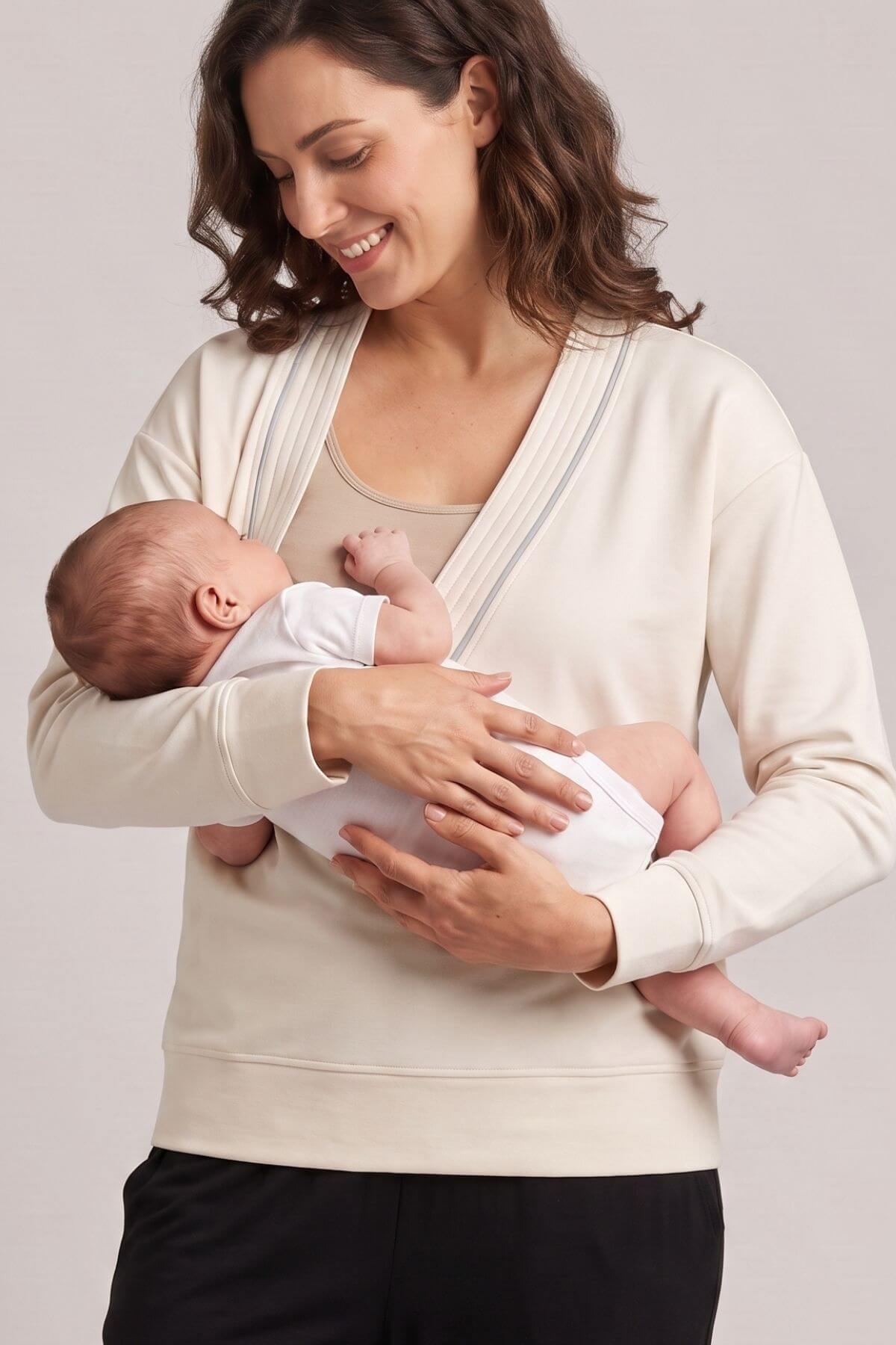 A woman wearing a cream-colored nursing tank top with a front baby wrap, holding a baby dressed in a white onesie. The background is a solid light gray studio backdrop.