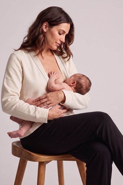 A woman sitting on a wooden stool breastfeeds a baby. She is wearing a cream-colored cardigan and black pants against a plain light gray studio background.