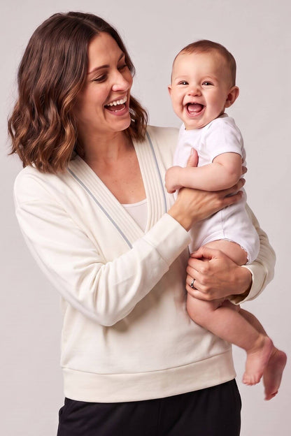 A woman holding a smiling baby, both wearing white clothing, photographed against a plain light gray studio background.