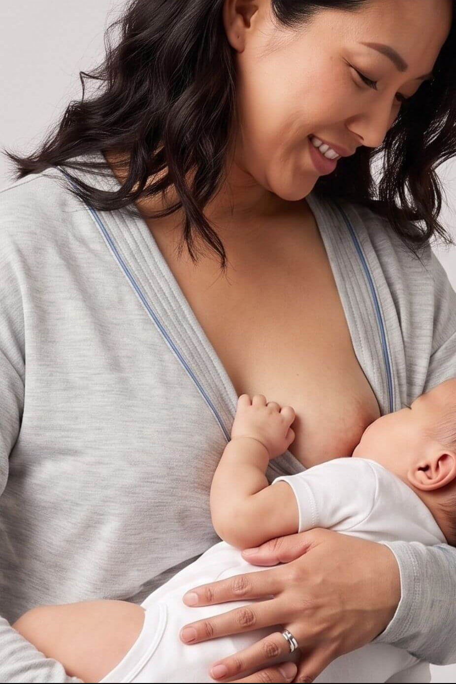 A woman wearing a light gray wrap top holds and breastfeeds a baby dressed in a white onesie against a plain light gray studio background.