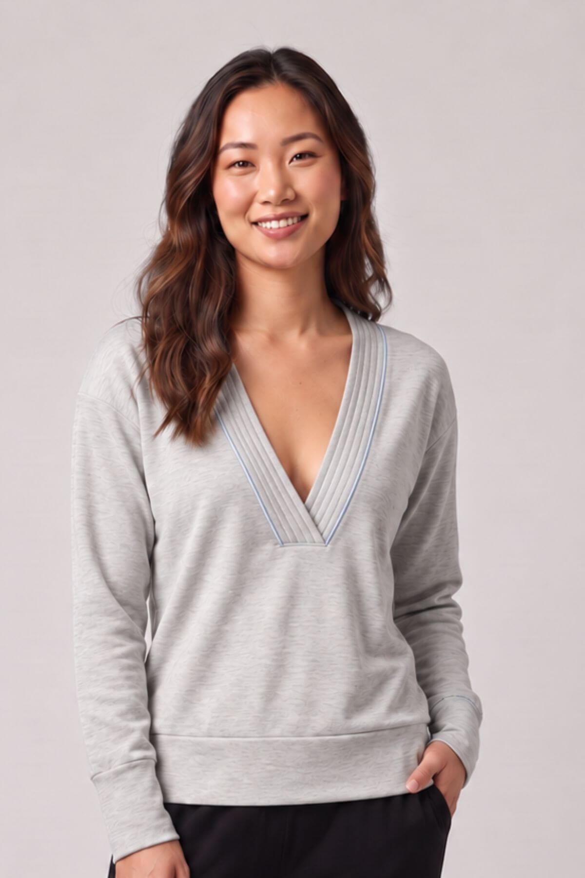 A woman with long brown hair smiling, wearing a light gray long-sleeve knit sweater with a V-neckline accented by thin blue stripes, set against a plain light gray studio background.