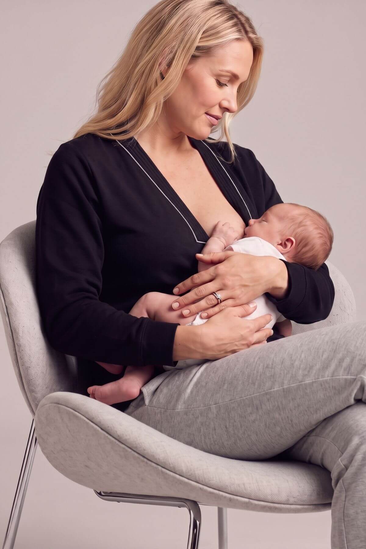 A woman in a black long-sleeve nursing top and gray sweatpants sits in a light gray upholstered chair, breastfeeding a baby wearing a white onesie..