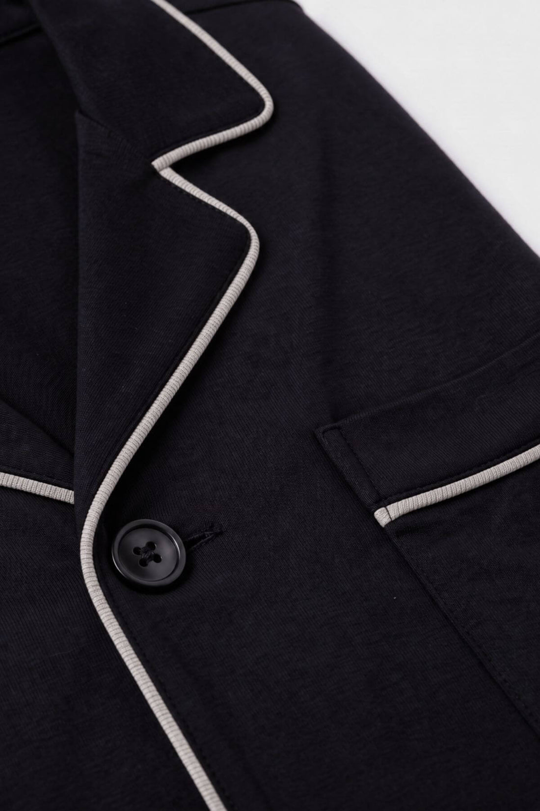 Close-up of a dark navy or black blazer with contrasting light gray piping along the lapel and pocket edges, featuring a black button.