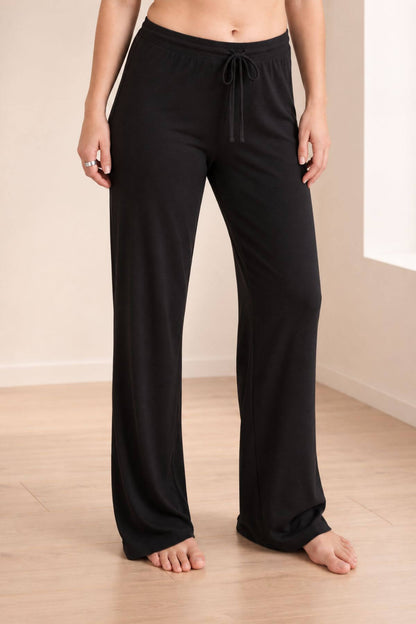 A person wearing black wide-leg lounge pants with a drawstring waist, standing barefoot on a light wood floor against a beige wall.