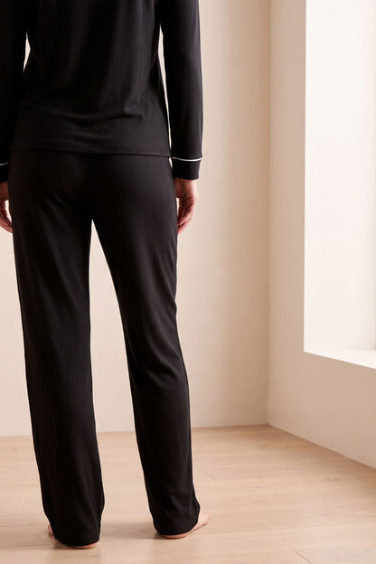 A person shown from the back wearing a black long-sleeve pajama top and matching black pants in a softly lit room with beige walls and light wood flooring. 