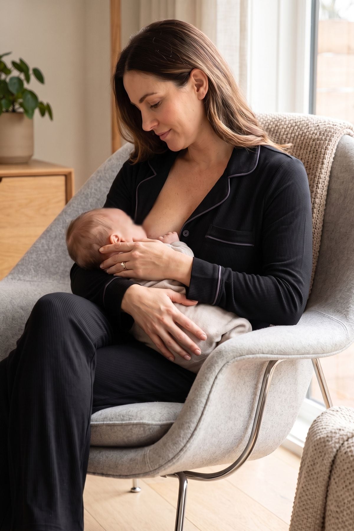 A woman in black pajamas sits in a light gray armchair, breastfeeding a baby wrapped in a light beige blanket in a softly lit room. 