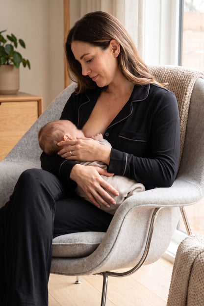 A woman in black pajamas sits in a light gray armchair, breastfeeding a baby wrapped in a light beige blanket in a softly lit room. 