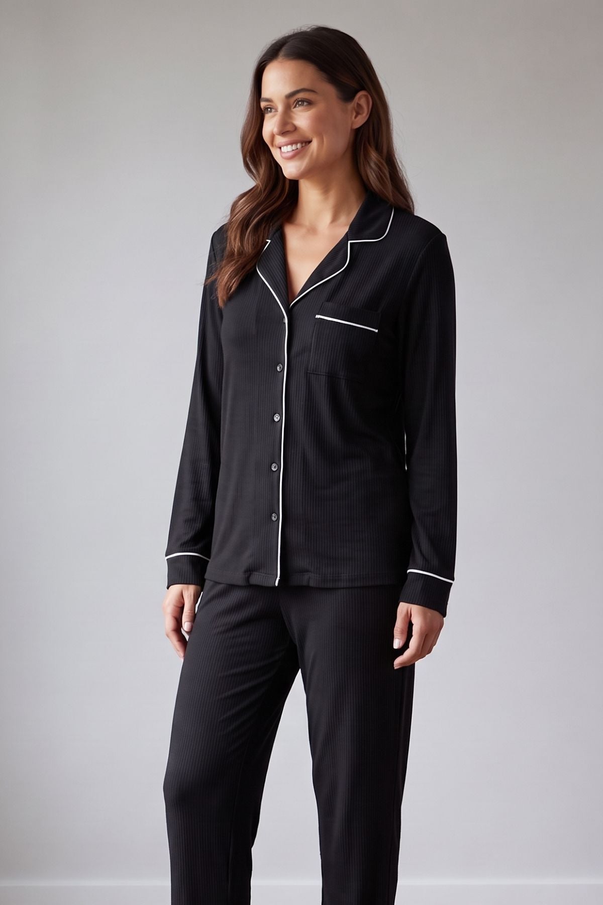 A woman modeling a black long-sleeve pajama shirt with white piping trim and matching black pajama pants against a light gray studio background. 