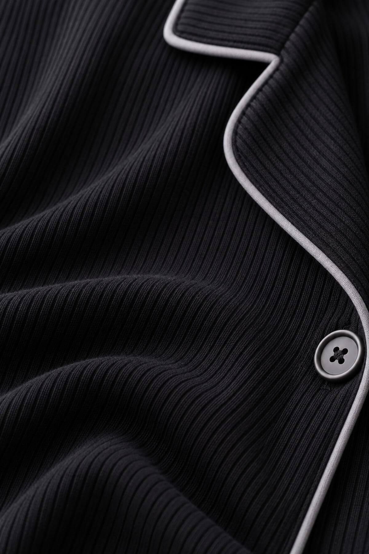 Close-up of a black ribbed knit garment, likely a cardigan or sweater, with contrasting light gray piping and a matching button.