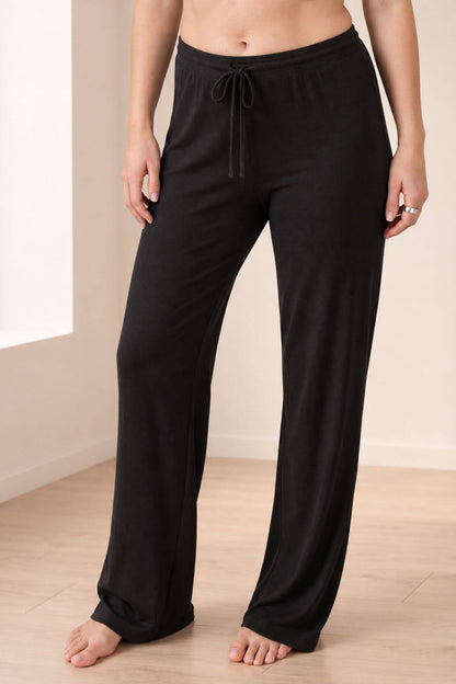 Black wide-leg lounge pants with an elastic drawstring waist, shown on a model standing barefoot in a brightly lit room with light wood floors.