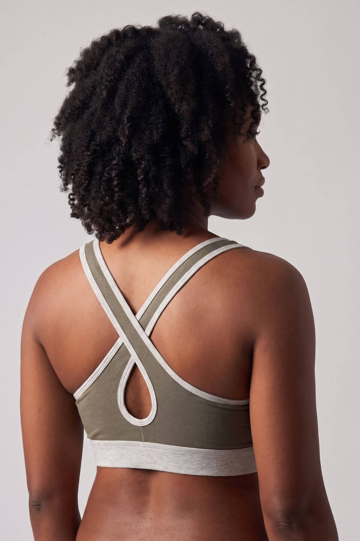 Rear view of a woman with curly black hair wearing an olive green and white sports bra with a crossover back strap design, set against a plain light gray studio background.