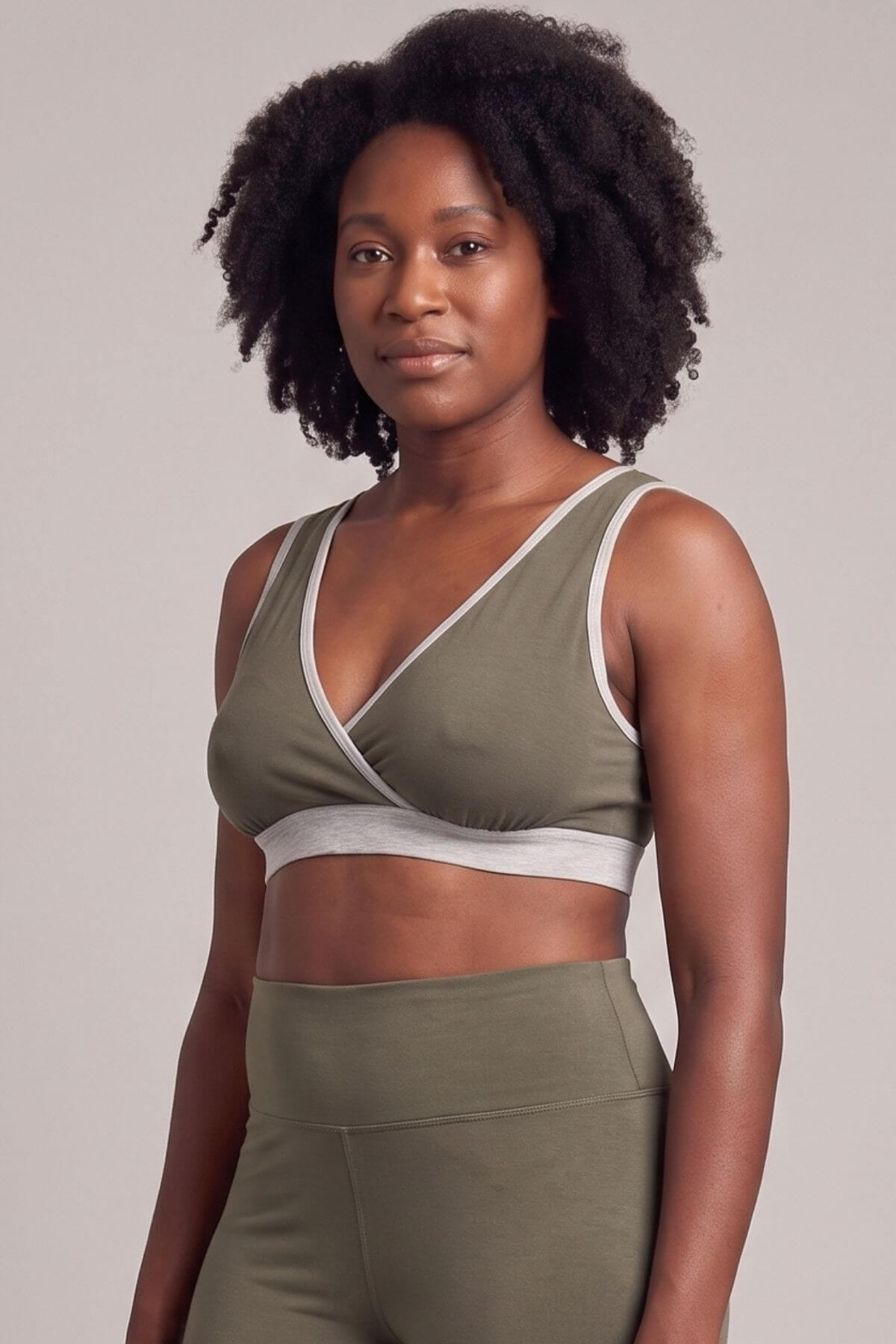 A woman modeling an olive green activewear set consisting of a crossover sports bra with white trim and matching high-waisted leggings, against a solid light gray background.