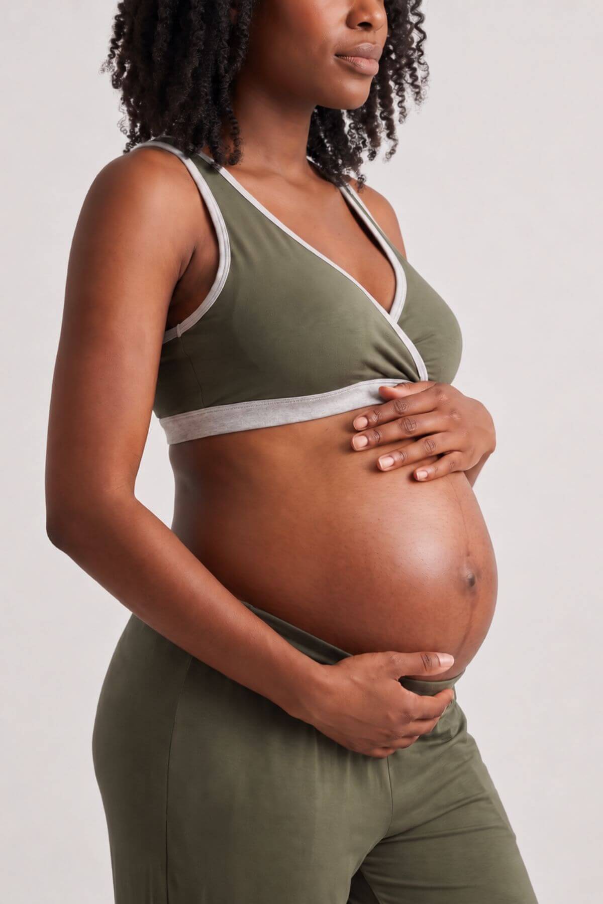 A pregnant woman wearing an olive green maternity sports bra and matching leggings, holding her baby bump against a light gray studio background.